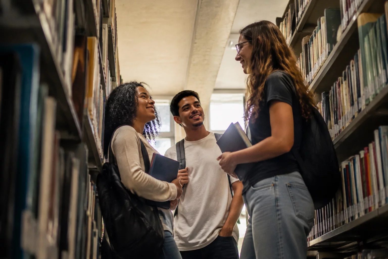 Estudantes conversando em pé entre as estantes de uma biblioteca